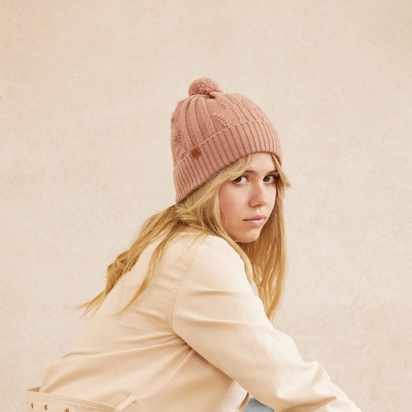 Woman wearing a blush pink beanie and beige jacket against a beige background
