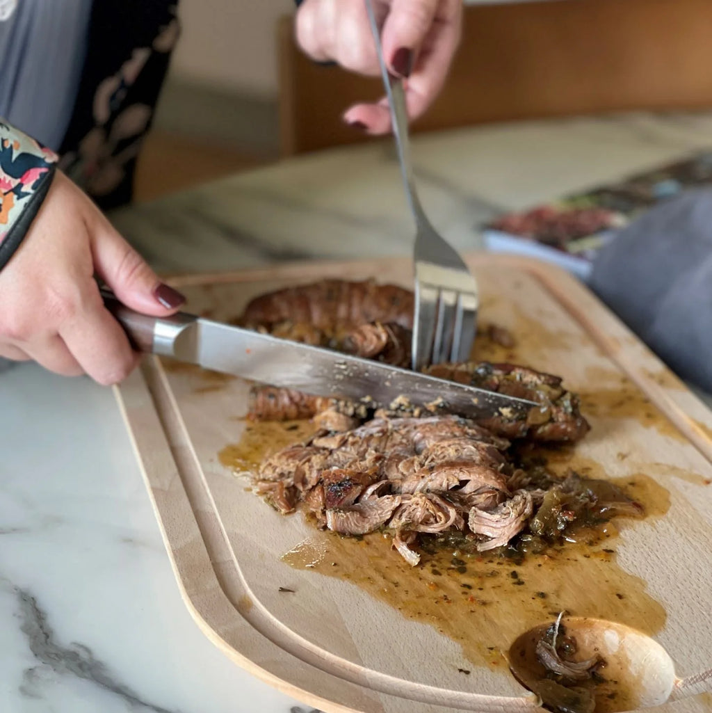 Person cutting into a piece of meat on a wooden board with a fork and knife.