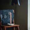 Pink candle in a white candle holder on a wooden chair with a dark floral-patterned cushion against a plain wall.