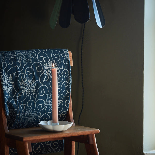 Pink candle in a white candle holder on a wooden chair with a dark floral-patterned cushion against a plain wall.