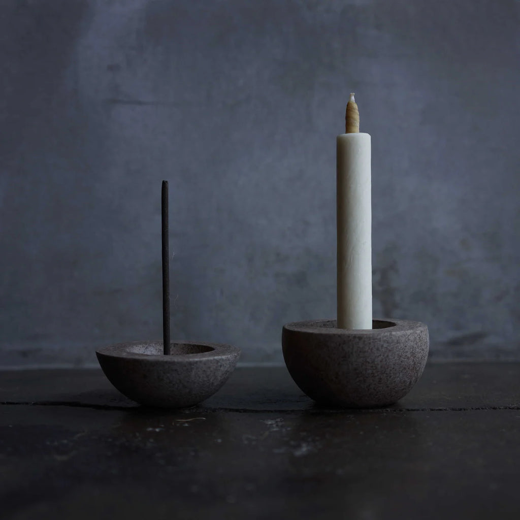 Two stone candle holders with a white candle and incense stick on a dark surface.