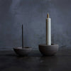 Two stone candle holders with a white candle and incense stick on a dark surface.