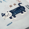A close-up of a white woven textile featuring a large embroidered cat design in navy and white, with small decorative motifs including windows, a bird, a fish, and partial text