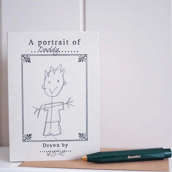 A square white card or print with the text 'A portrait of Daddy' and a simple hand-drawn sketch of a child, placed on a tan surface next to a dark green pen with gold lettering.
