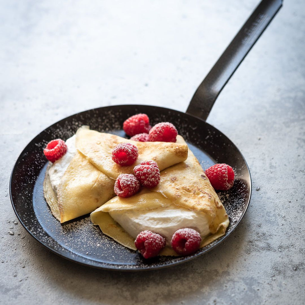 Two folded crepes on a frying pan with raspberries on a light gray background