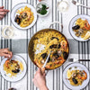 Overhead view of a seafood paella spread on a striped tablecloth, with a large pan of yellow rice, shrimp, mussels, and fish in the center and several white plates being served by visible hands.