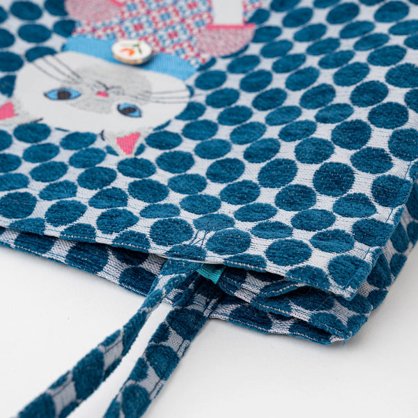 A close-up of a fabric tote bag with a blue and white polka dot pattern, featuring a white cat face patch and a pink patterned fabric accent on the top left.