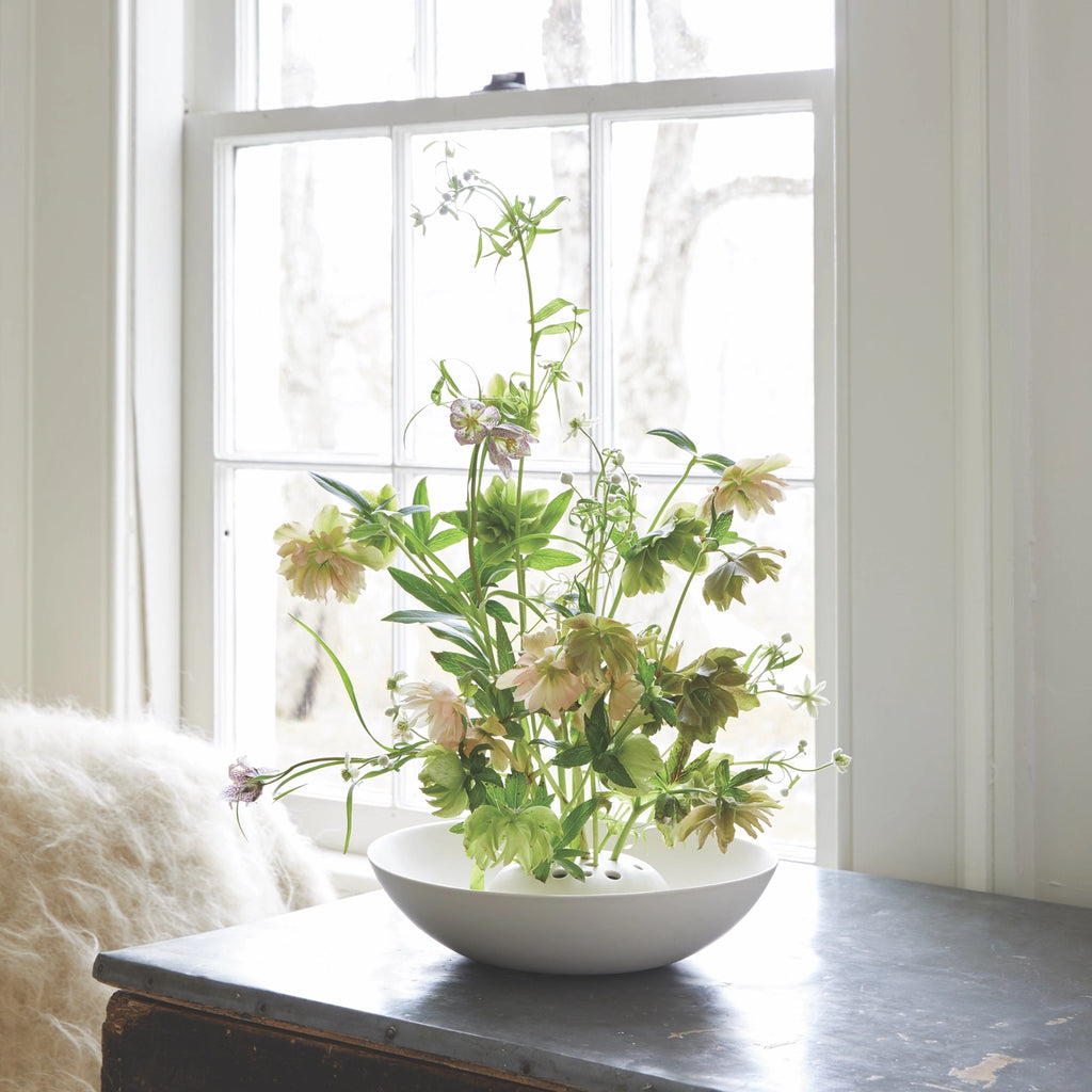 Bouquet of flowers in a white bowl on a table with a window in the background