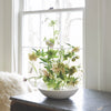 Bouquet of flowers in a white bowl on a table with a window in the background