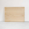 A rectangular light wood cutting board with visible natural grain patterns, standing upright against a plain white wall on a light gray surface.