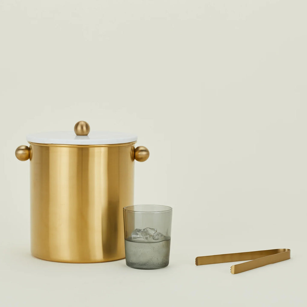 A brass finish ice tong with a lid, alongside a clear glass and a metal straw.