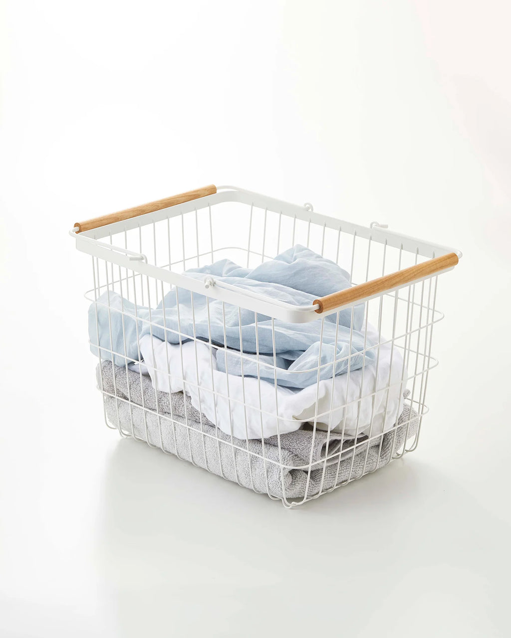 White wire laundry basket with wooden handles filled with clothes on a white background