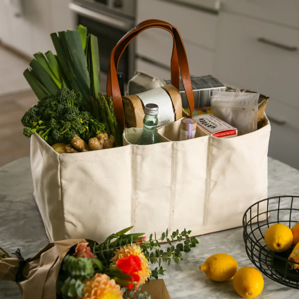 A beige fabric tote bag with a brown leather handles attached to the side, filled with various items veggies and beverages.