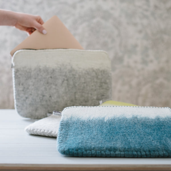 A hand is placing a beige laptop into a soft shell laptop case which is white on top and gray at the bottom with a woolen texture. Other laptop cases in gray, blue, and yellow are visible in the background.