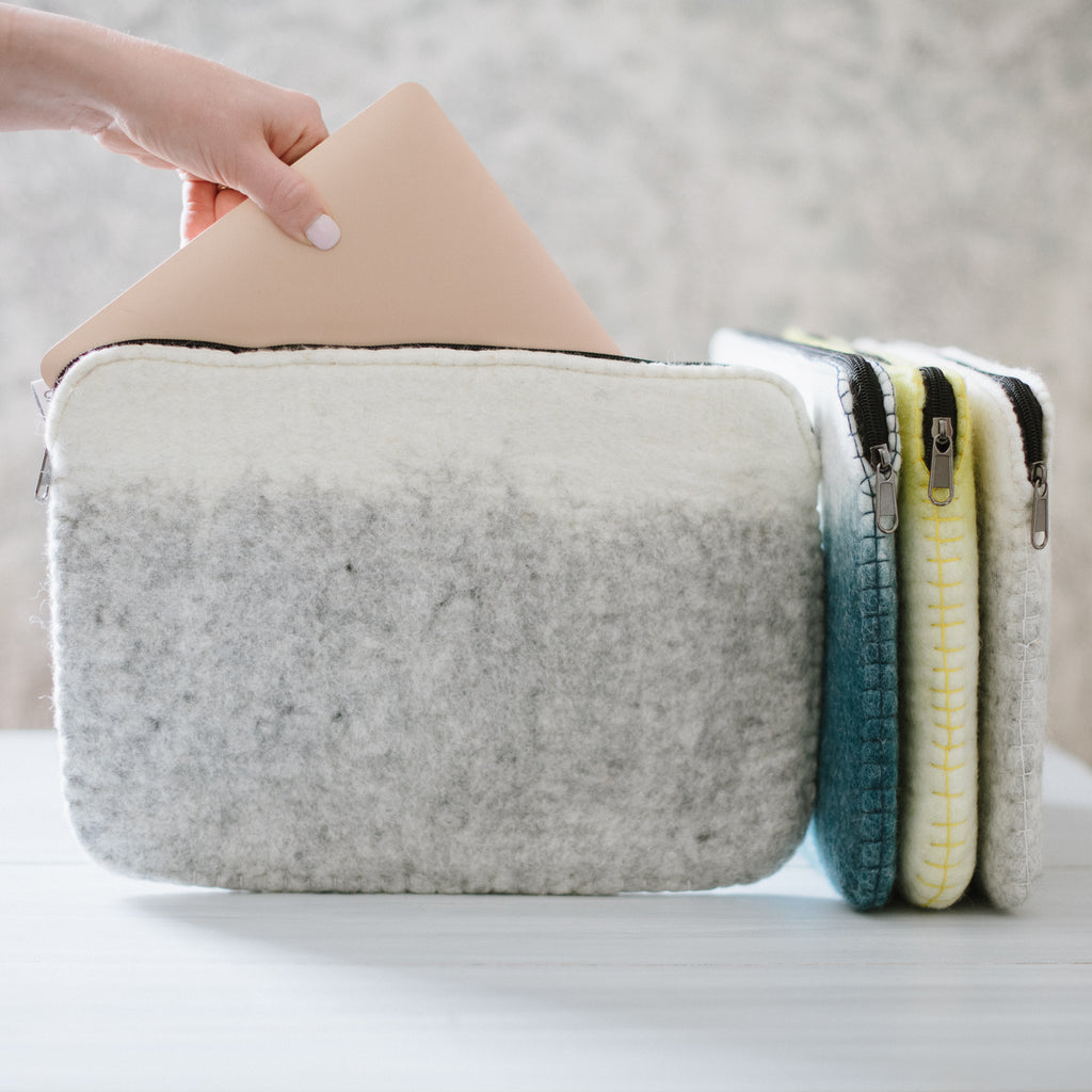 A hand is placing a beige laptop into a soft shell laptop case which is white on top and gray at the bottom with a woolen texture. Other laptop cases in gray, blue, and yellow are visible in the background.