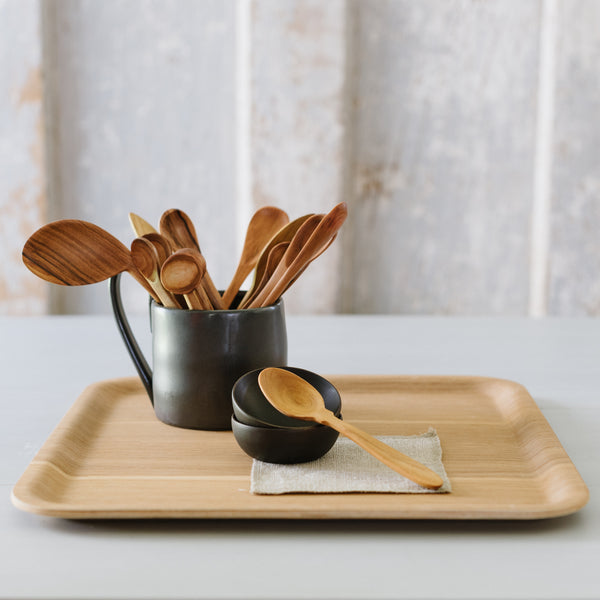 Wooden spoons in a black coffee mug on a wooden tray. One spoon is resting on 2 small black wooden bowls with a grey cocktail napkin underneath. 