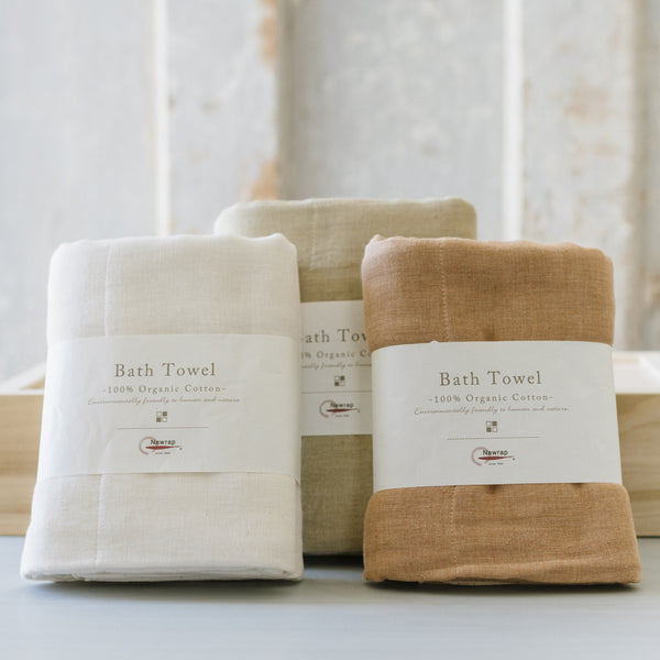 Three folded organic cotton bath towels in beige, brown, and white colors, displayed on a flat surface with a textured background.