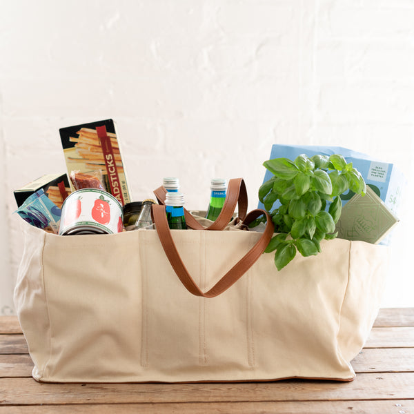 A beige fabric tote bag with a brown leather handle attached to the side, filled with various items such as food and bottled water.