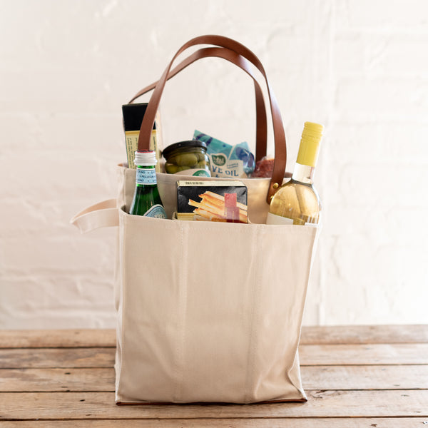 A beige fabric tote bag with a brown leather handle attached to the side, filled with various items such as food and bottled water, side view.