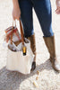 A person holding a beige fabric tote bag with a brown leather handle attached to the side, filled with various items such as a blanket and wine.