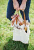 A person holding a beige fabric tote bag with a brown leather handle attached to the side, filled with various items such as a blanket and wine.