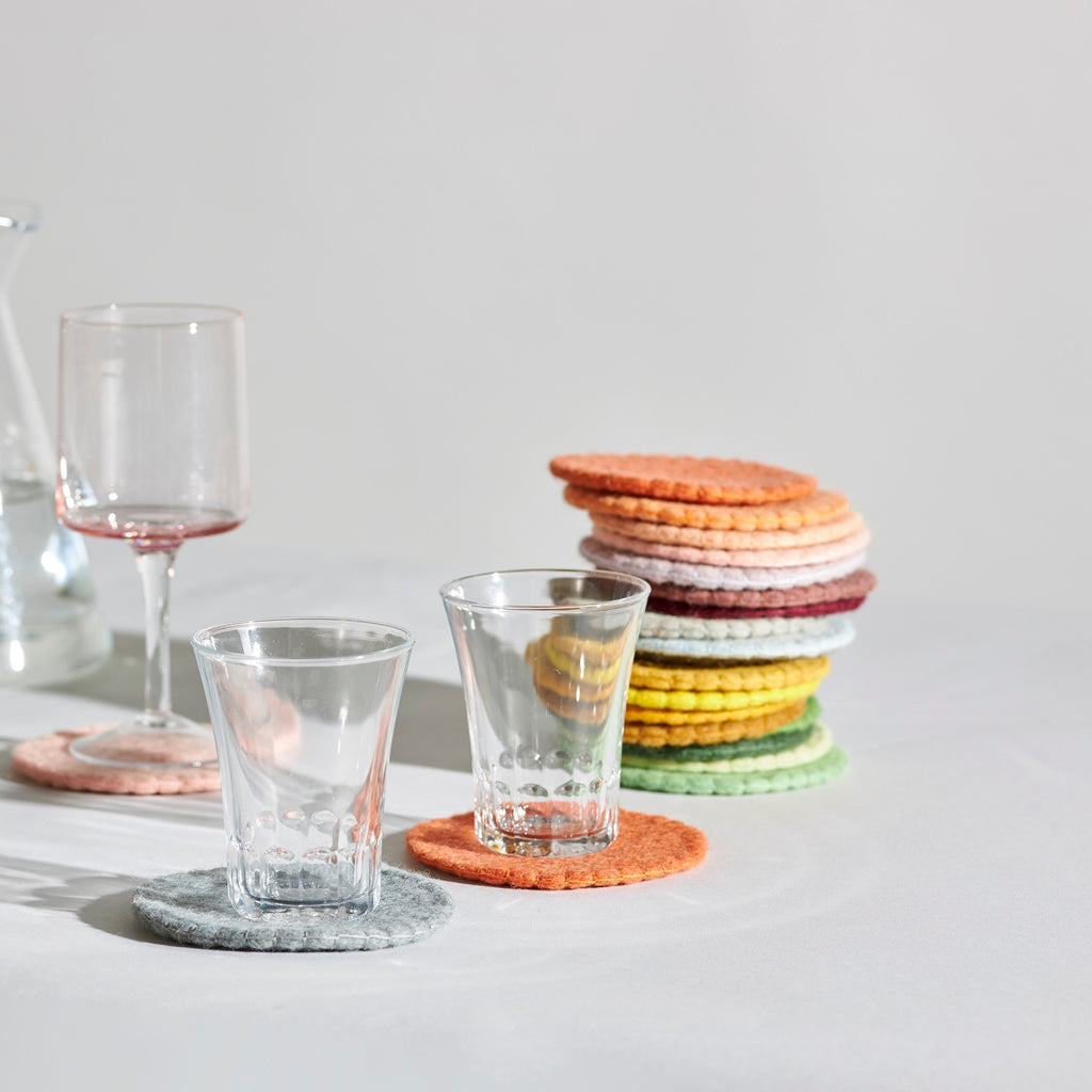 Handmade wool coasters in various natural colors placed on a white surface with two glasses of different sizes.